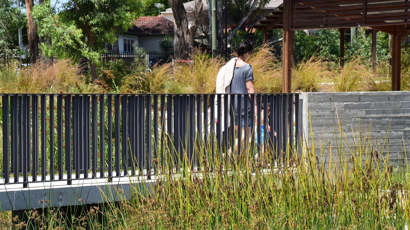 A person with short dark hair, wearing a gray shirt and blue shorts, is seen from the back walking across a bridge with a black railing over the grassy area of Blacktown Showground. A towel is draped over their shoulder. Trees and vegetation are in the background, along with buildings.