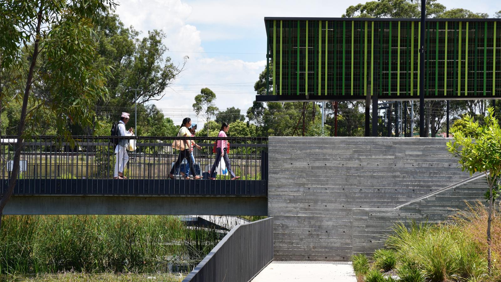 Four people walk across a modern footbridge over a wetland area with tall grass at Blacktown Showground. The landscape features greenery and a modern building with green vertical panels in the background. The sky is partly cloudy.