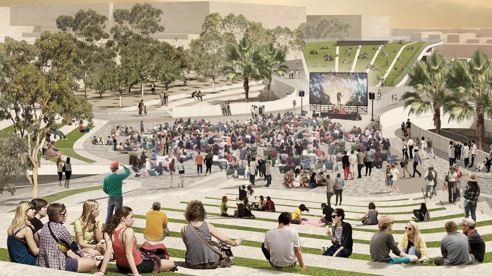 Outdoor amphitheater scene with people seated on tiered steps watching a large screen showing a concert. Trees, greenery, and buildings surround the area, including Cato St nearby. Many viewers are sitting on the ground while others are standing, creating a lively and social atmosphere.