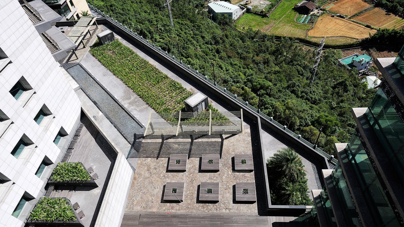 Aerial view of an elevated, terraced garden and outdoor space attached to a modern building. The area features a mix of stone-paved surfaces, garden plots, wooden decking, and seating arrangements. In the background, lush green forests and farmland are visible near Chang Gung Hospital.