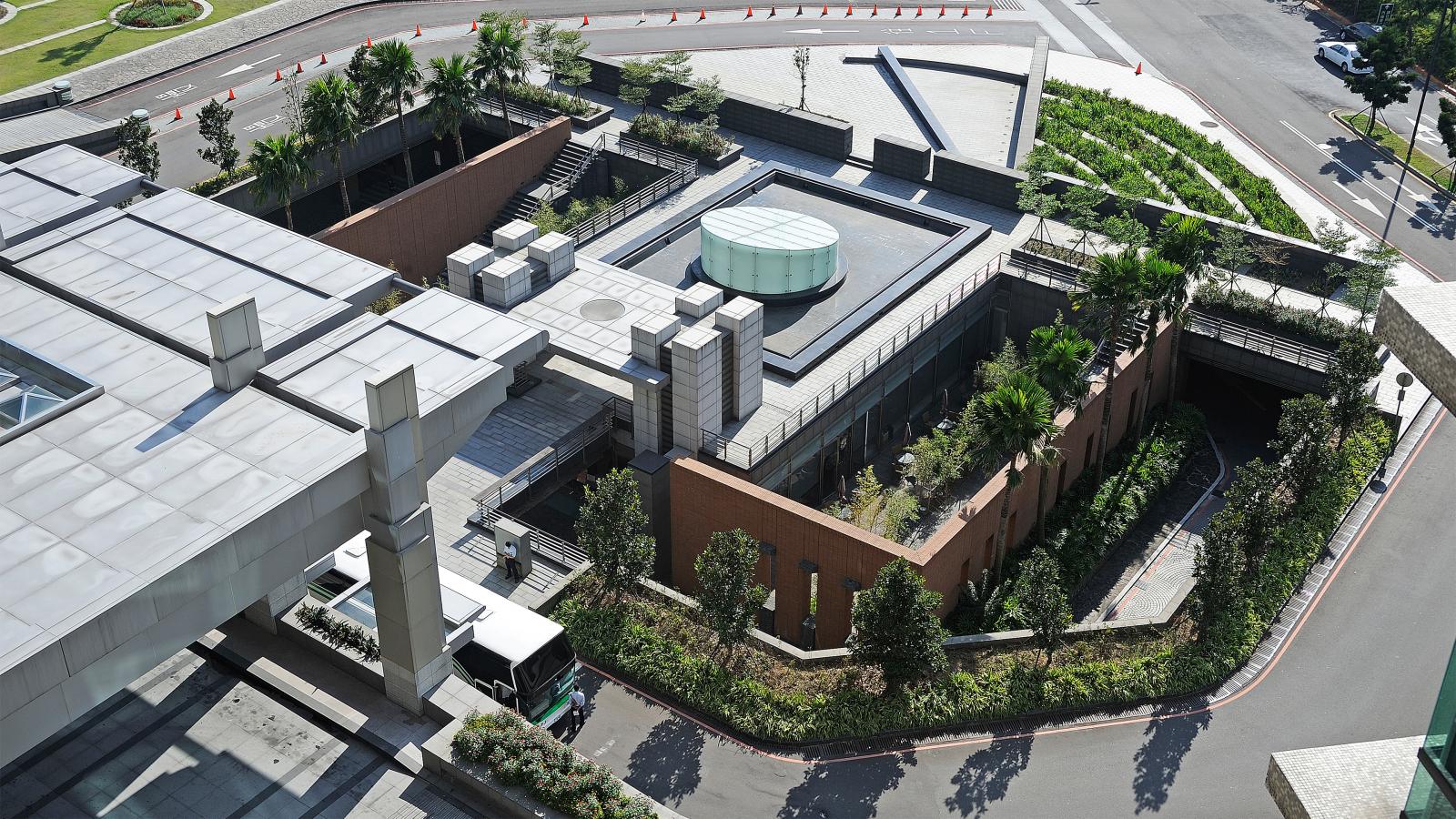 Aerial view of a modern building complex, reminiscent of Chang Gung Hospital, featuring a mix of concrete, glass, and greenery. The design includes rooftop gardens, a circular skylight, and landscaped pathways. The surrounding area has roads and parked vehicles against a backdrop of urban elements and lush greenery.