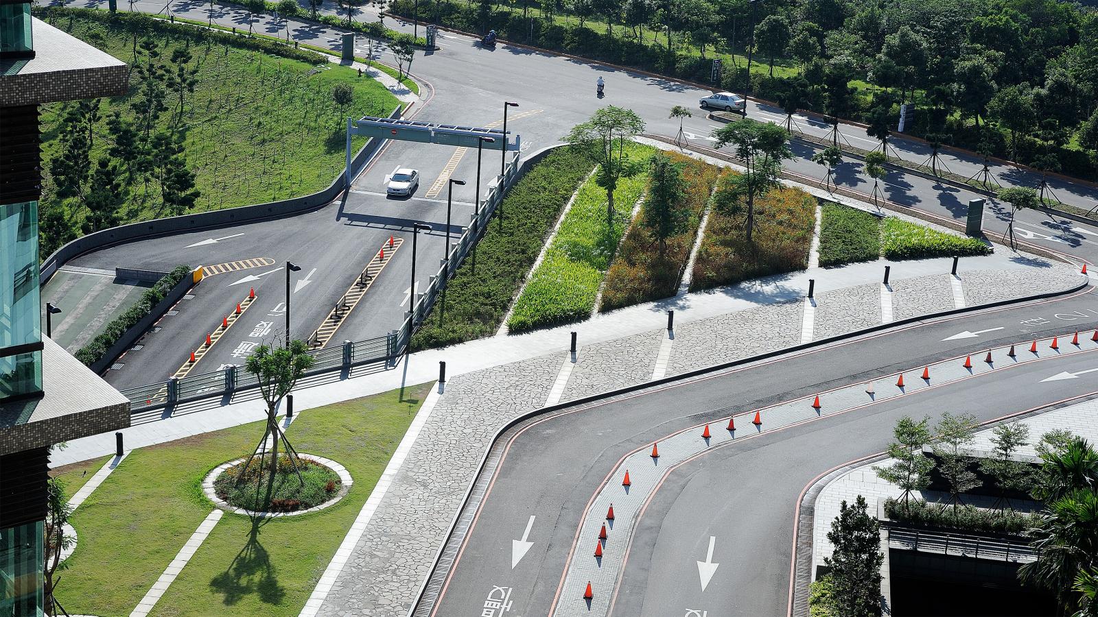 Aerial view of a modern road leading to Chang Gung Hospital, with a secured entrance featuring a white car arriving at the gate. The well-maintained road includes landscaped areas with grass and trees, orange traffic cones, and pathways, set against a backdrop of greenery and trees.