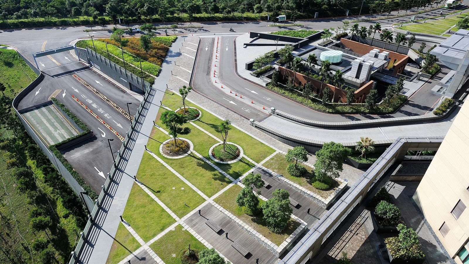 Aerial view of a modern green landscape featuring neatly trimmed bushes, trees, and grass areas. Paved walkways and roads with marked lanes weave through the scene. Buildings, including Chang Gung Hospital with rooftop gardens, are visible alongside lush greenery and parking areas.