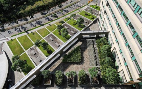 Aerial view of a modern building with green rooftop terraces and landscaped gardens. The terraces feature trees, bushes, and seating areas. Adjacent to the building is a road with cars, separated by a line of trees and a sidewalk, reminiscent of the layout at Chang Gung Hospital.