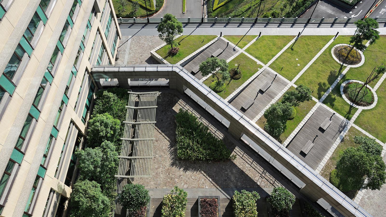 An aerial view of a landscaped urban area beside Chang Gung Hospital reveals green lawns, trees, and wooden benches. A long pedestrian bridge, supported by metal pillars, spans through the area, connecting different sections of the space seamlessly.