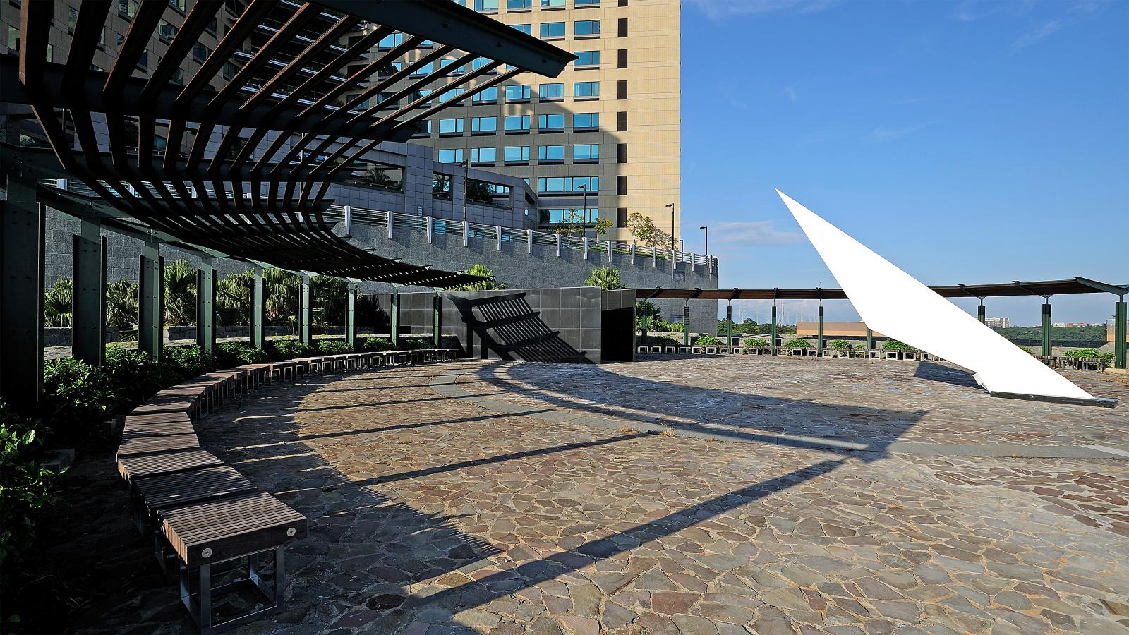 A modern outdoor plaza with stone flooring and curved wooden benches. A large, angled white sculpture stands prominently. Overhead, a series of interconnected black beams create a partial shade structure. In the background, Chang Gung Hospital rises tall against the clear blue sky.