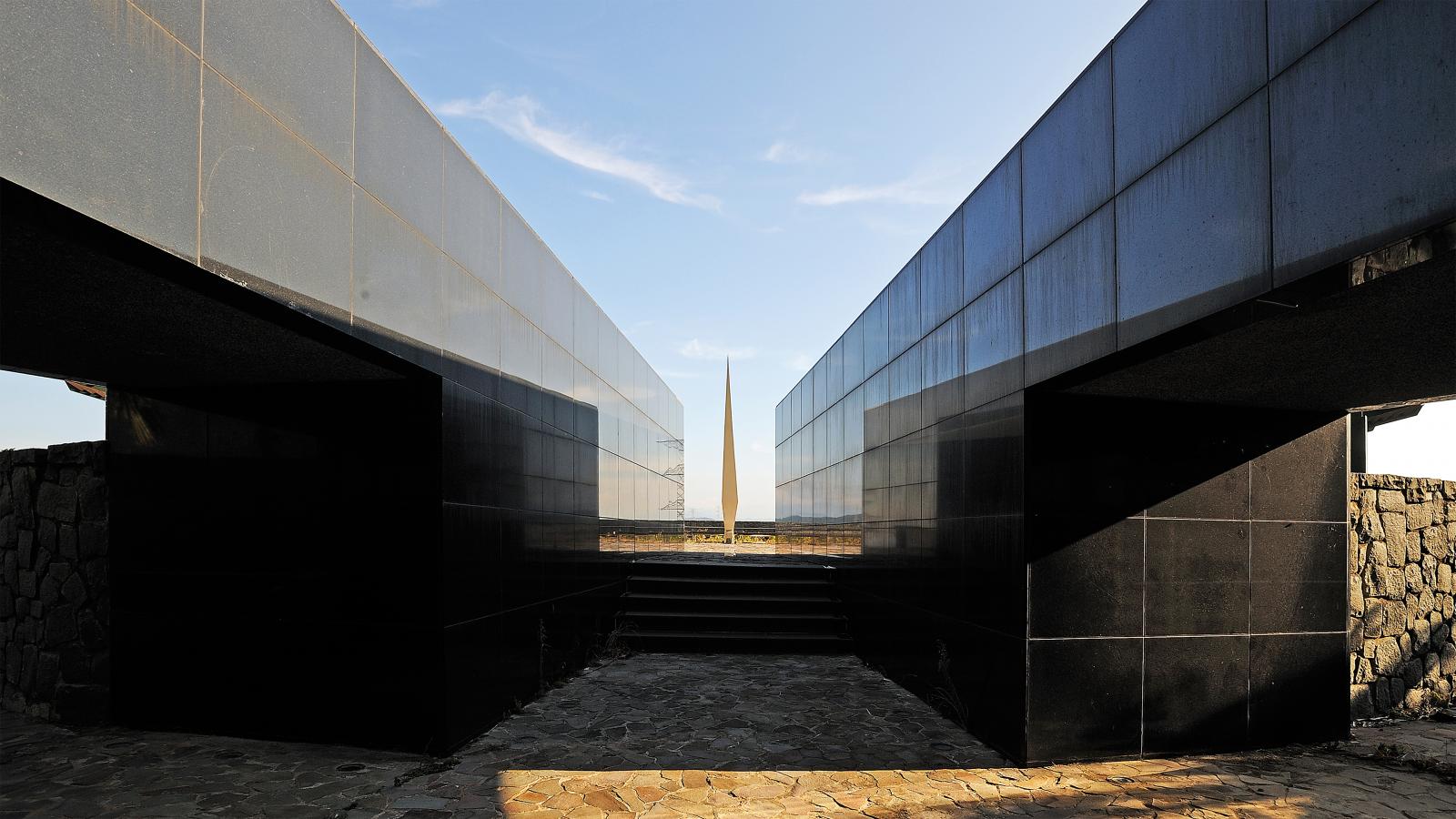 A symmetrical image shows a pathway leading between two tall, sleek, dark stone walls towards a distant, tall obelisk under a blue sky. The ground is paved with irregular stone tiles, and the mirrored walls reflect the pathway and sky, evoking the meticulous planning of Chang Gung Hospital.