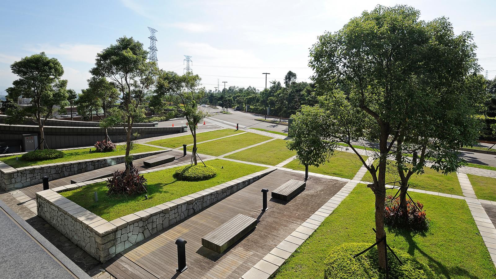 Public park with neatly trimmed grass, paved pathways, wooden benches, and symmetrically planted trees. The area is bordered by a stone wall, and a road leading to Chang Gung Hospital with electric pylons is visible in the background under a clear sky.