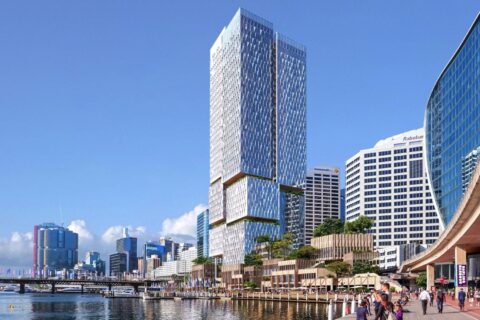 A modern city skyline featuring a tall, reflective skyscraper with unique stacked sections dominates the scene. Below, the bustling Cockle Bay Promenade has people walking and enjoying the view. Other high-rise buildings are visible under a clear blue sky at Cockle Bay Park.