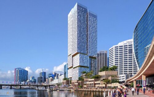 A modern city skyline featuring a tall, reflective skyscraper with unique stacked sections dominates the scene. Below, the bustling Cockle Bay Promenade has people walking and enjoying the view. Other high-rise buildings are visible under a clear blue sky at Cockle Bay Park.