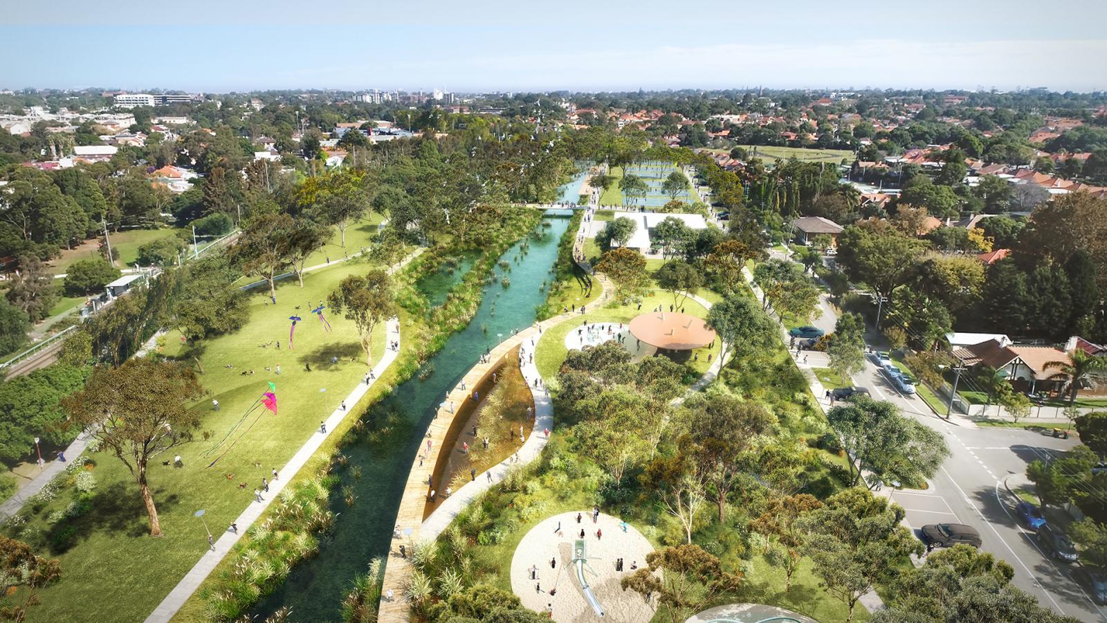 Aerial view of GreenWay park with a winding river flowing through it. Walking paths, small bridges, and shaded seating areas are visible. People are enjoying various activities, and there are residential buildings and trees in the background.