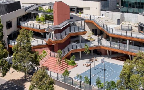 Docklands Primary School's modern multi-level building features terraces with plants and outdoor seating. A red central stairway connects the levels. The ground area includes a blue basketball court surrounded by trees, with adjacent seating and pathways.