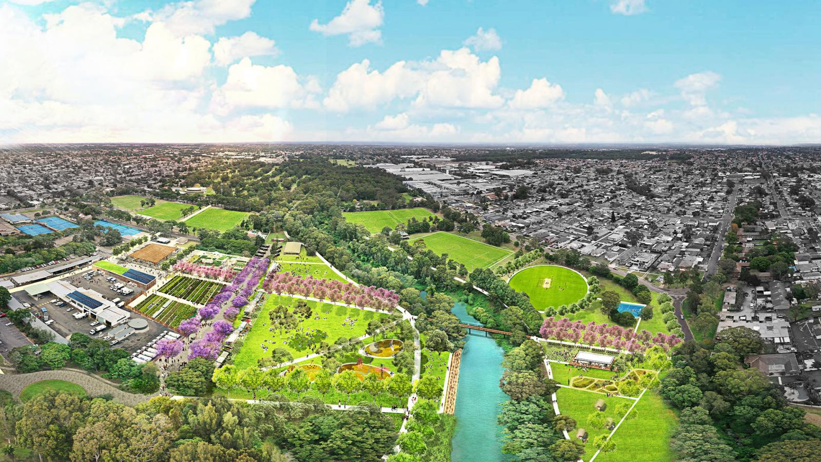 Aerial view of a vibrant urban park with green spaces, sports fields, pathways, and clusters of trees and flowers along the scenic Duck River. The park, part of an extensive masterplan, is bordered by residential neighborhoods on one side and industrial buildings on the other under a partly cloudy sky.