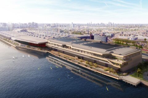 Aerial view of the impressive Excel London, a large modern waterfront building with multiple levels and terraces, situated along a river. Several small boats are in the water nearby. The background features a sprawling cityscape with various buildings and infrastructure under Stage 3 development. The sky is clear and blue.