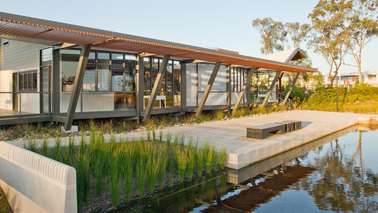 A modern single-story building with large windows and a mix of wood and metal elements sits next to a reflecting pond in Fairwater Community Park. The structure features angled supports and a terrace with a rectangular wooden bench. Trees and shrubs surround the property, enhancing its serene setting.