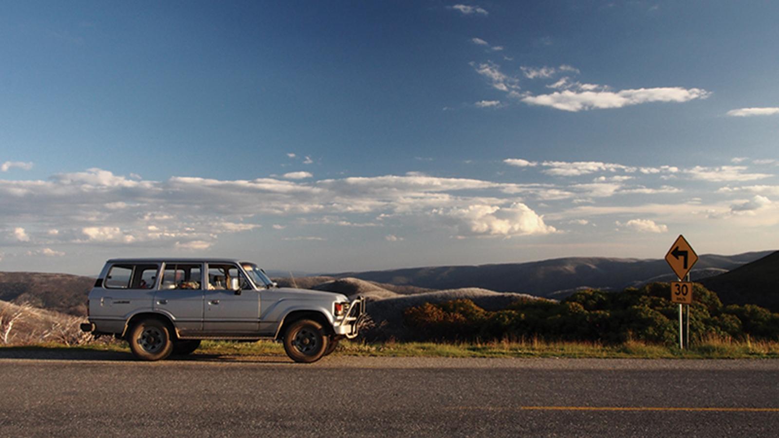 An older model silver SUV is parked on the side of a road with a mountainous landscape in the background. The sky is partly cloudy, and there is a yellow road sign indicating a right curve with a speed limit of 30 mph. This scenic route could be part of the Alpine Crossing to Falls to Hotham.