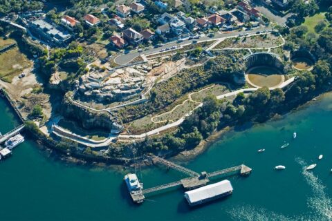 A scenic aerial view of a coastal area featuring a winding pathway through a hilly landscape. Residential houses are clustered near the top left, with boats anchored in the blue-green water on the bottom right. The former BP site has been transformed into a park, and a pier extends into the water with a large docked structure.