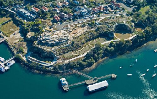 A scenic aerial view of a coastal area featuring a winding pathway through a hilly landscape. Residential houses are clustered near the top left, with boats anchored in the blue-green water on the bottom right. The former BP site has been transformed into a park, and a pier extends into the water with a large docked structure.