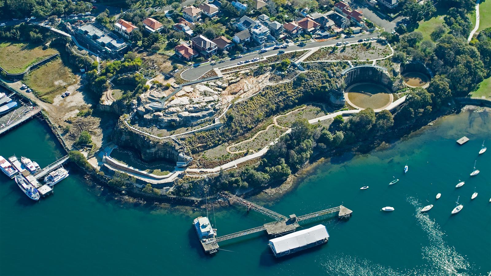 A scenic aerial view of a coastal area featuring a winding pathway through a hilly landscape. Residential houses are clustered near the top left, with boats anchored in the blue-green water on the bottom right. The former BP site has been transformed into a park, and a pier extends into the water with a large docked structure.