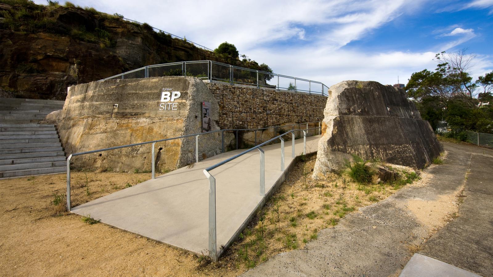 A concrete ramp and metal railing lead to a viewing platform integrated into a rocky landscape at the Former BP Site Park. The platform, surrounded by wire mesh fencing, features the initials "BP SIT" painted on a large rock. Stairs are on the left, and trees are visible in the background.