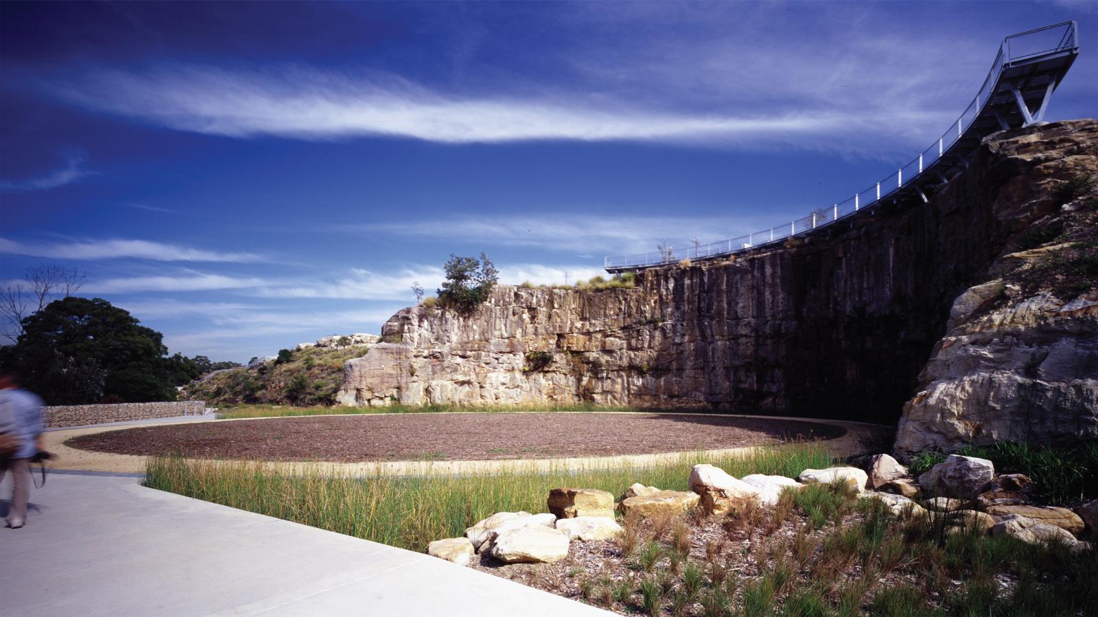 A path leads to a circular, sandy area surrounded by tall rocks at Former BP Site Park. The rocky cliff, topped by a modern walkway with railings, dominates the scene under a mostly clear sky with light clouds. Vegetation is sparse, with grass and a few bushes.