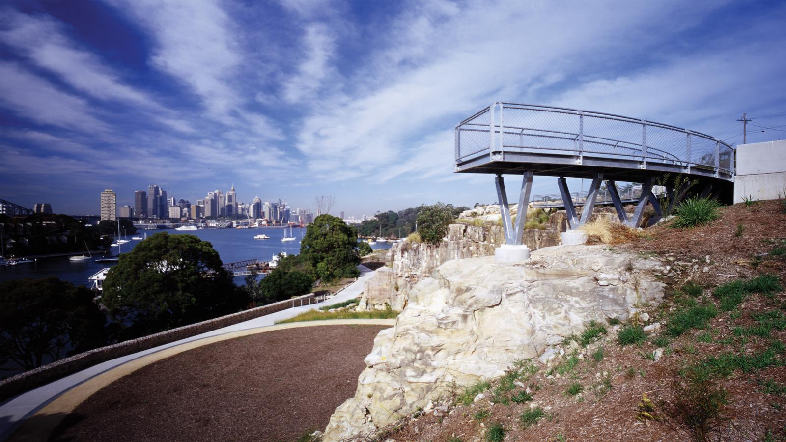 A scenic view of a modern lookout platform extending over a cliffside, offering a panoramic vista of a city skyline in the distance. Below, a winding pathway weaves between greenery and rocky terrain in this park, once a former BP site, under a partly cloudy blue sky.