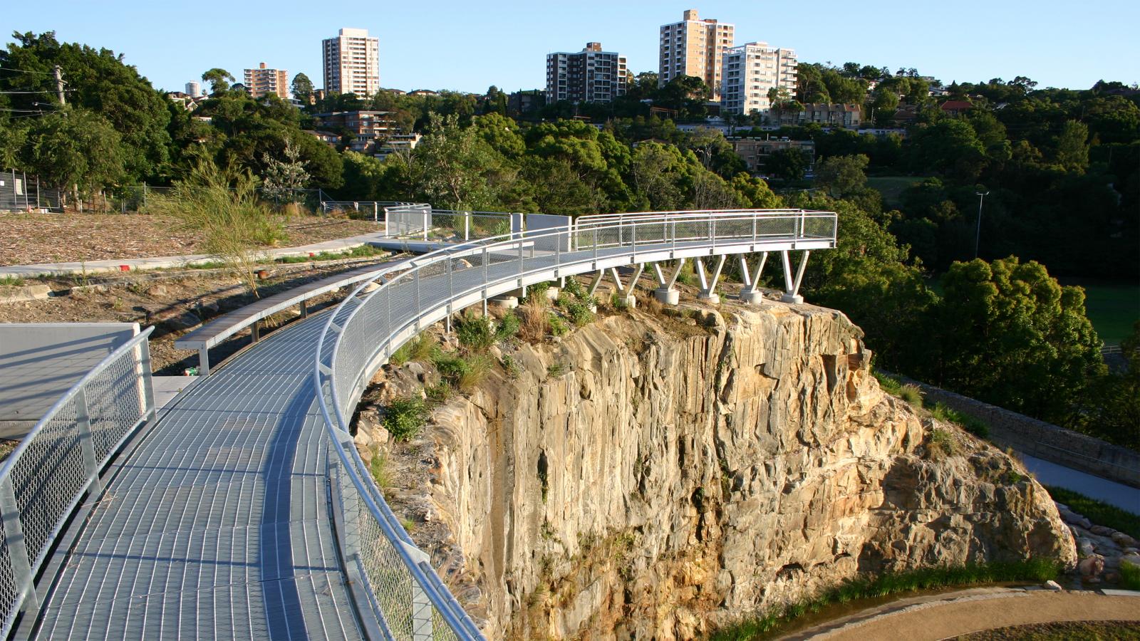 A metal pedestrian walkway curves along the edge of a rocky cliff, supported by sturdy beams. The cliff overlooks BP Site Park, with lush green areas and tall buildings in the background under a clear blue sky. Trees and vegetation are scattered throughout this beautifully reclaimed former BP site.
