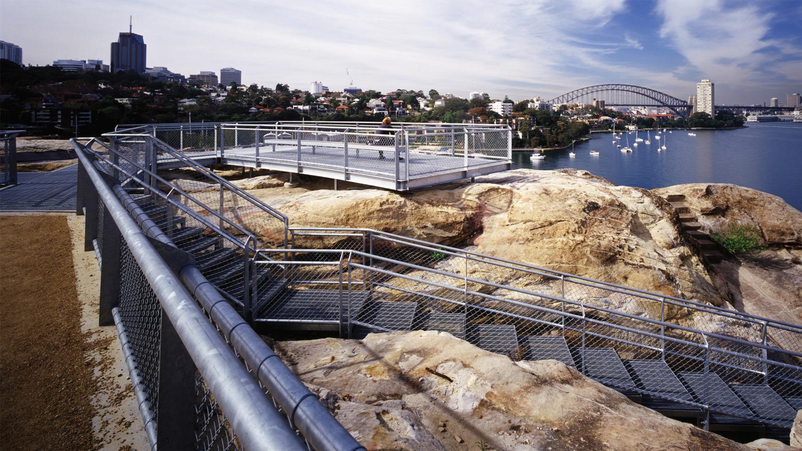 View from a rocky coastal area with multiple metal railings and platforms, overlooking a bay. The space, once a former BP site, features a cityscape and a prominent arched bridge in the distance, with a few boats anchored in the water under a partly cloudy sky.