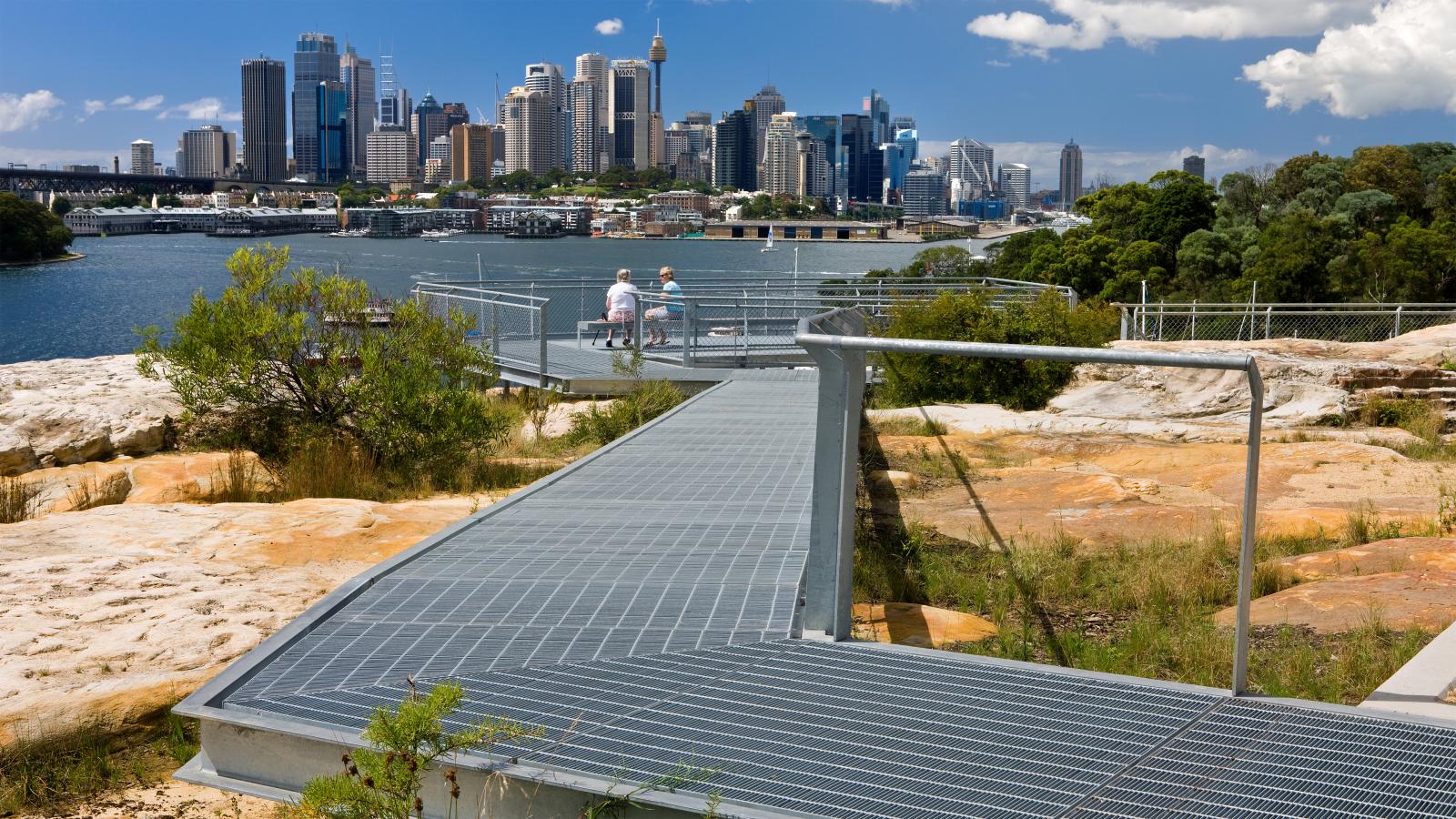 A metal walkway in the park leads to a lookout area where two people are seated on a bench, enjoying a scenic view of the city's skyline across a body of water. The landscape around the former BP site features rocky terrain, greenery, and clear blue sky with a few clouds.