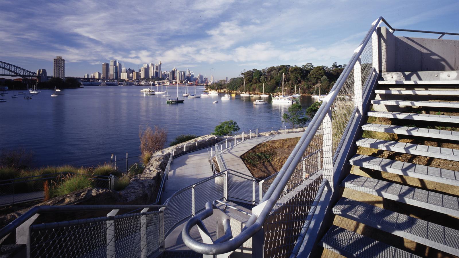 A view of Former BP Site Park's waterfront path and staircase, overlooking a harbor filled with anchored boats. A city skyline with tall buildings is visible in the background, under a partly cloudy sky. The scene is framed by modern railings and well-kept landscaping.