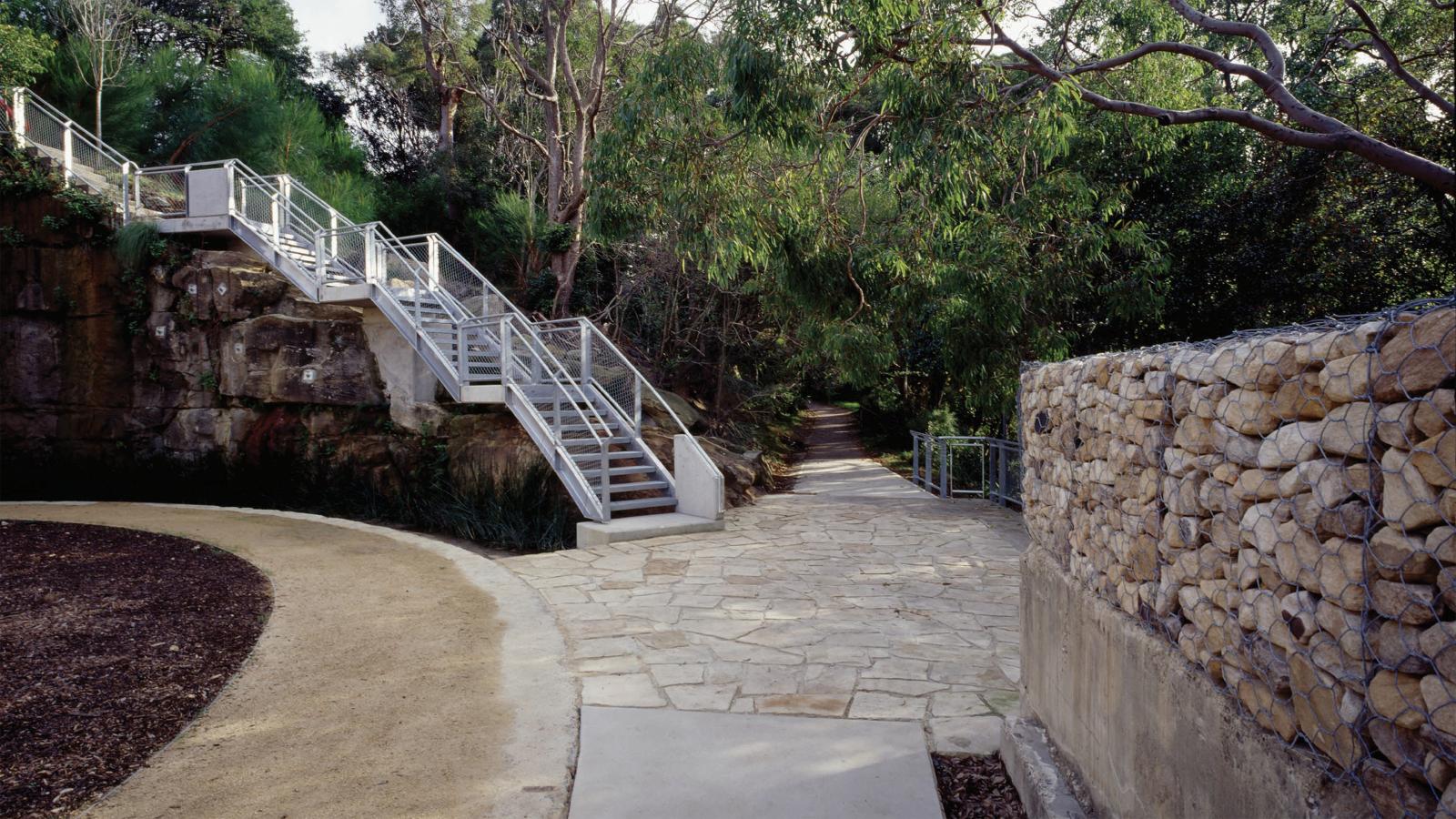 A stone pathway in the park leads to a metal staircase ascending a rocky hill. The path is bordered by a curved dirt section on one side and a stone wall with metal fencing on the other. Dense greenery surrounds the area, with trees overhanging the walkway, transforming this former BP site into an urban oasis.