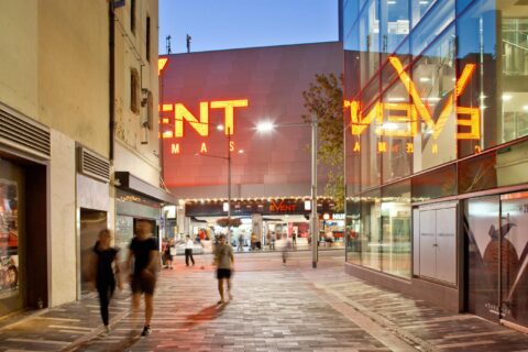 A modern urban street scene at dusk, with people walking past brightly lit buildings along George St Laneways. Large neon signage for "Event Cinemas" glows in the background, reflecting off nearby glass walls. The scene is lively, featuring a blend of architecture and commercial activity.