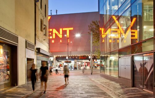 A modern urban street scene at dusk, with people walking past brightly lit buildings along George St Laneways. Large neon signage for "Event Cinemas" glows in the background, reflecting off nearby glass walls. The scene is lively, featuring a blend of architecture and commercial activity.