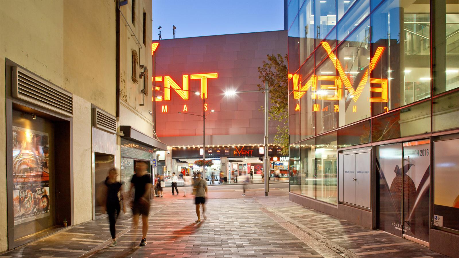 A modern urban street scene at dusk, with people walking past brightly lit buildings along George St Laneways. Large neon signage for "Event Cinemas" glows in the background, reflecting off nearby glass walls. The scene is lively, featuring a blend of architecture and commercial activity.