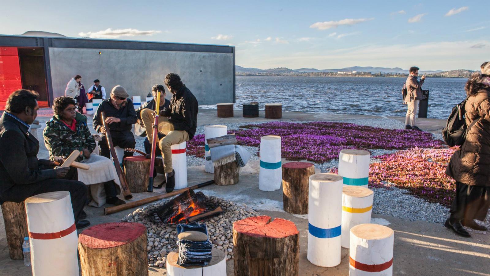 A group of people sits around a fire pit by the water in Glenorchy, playing musical instruments on colorful, cylindrical stools. In the background, others stroll along the shoreline near an artful purple flower arrangement. The clear sky adds to the serene atmosphere of this sculpture park setting.