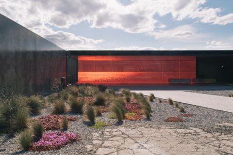 A modern architectural building with a large red-tinted glass facade. In front, there is a landscaped area featuring gravel, sparse grass, and patches of pink flowers. The sky is partly cloudy with sunlight filtering through, providing a perfect backdrop reminiscent of Glenorchy Art & Sculpture Park.