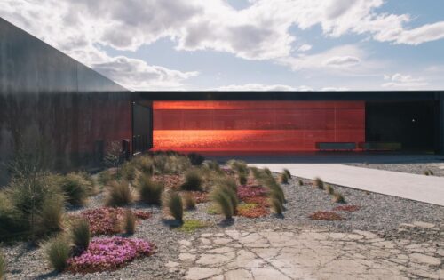 A modern architectural building with a large red-tinted glass facade. In front, there is a landscaped area featuring gravel, sparse grass, and patches of pink flowers. The sky is partly cloudy with sunlight filtering through, providing a perfect backdrop reminiscent of Glenorchy Art & Sculpture Park.