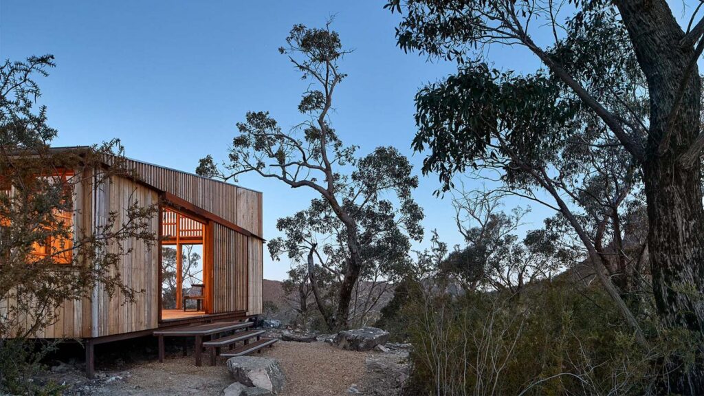 A small wooden cabin with large windows is situated in a remote, rocky landscape surrounded by tall trees and bushes along the Grampians Peaks Trail. The interior light illuminates the cabin's warm wooden panels, contrasting with the dusky blue sky and natural surroundings of Gariwerd.