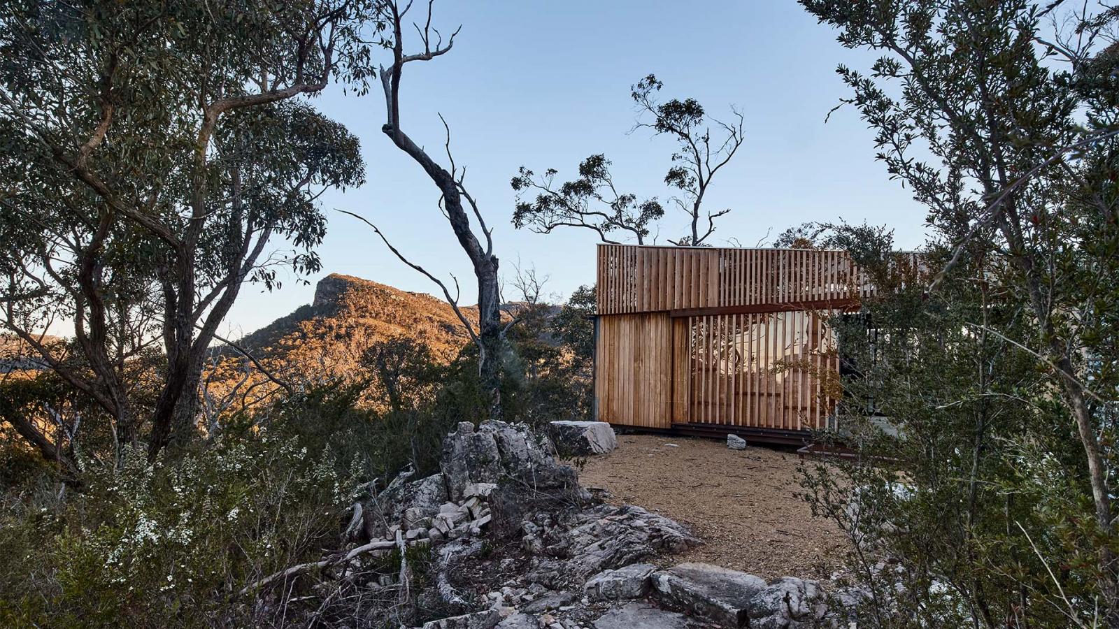 A modern wooden cabin is situated amidst the rocky, forested landscape of Gariwerd. Tall trees with sparse foliage surround the structure. The sun is setting, casting a warm glow on the nearby mountain peak along the Grampians Peaks Trail and creating long shadows. The sky is clear and blue.