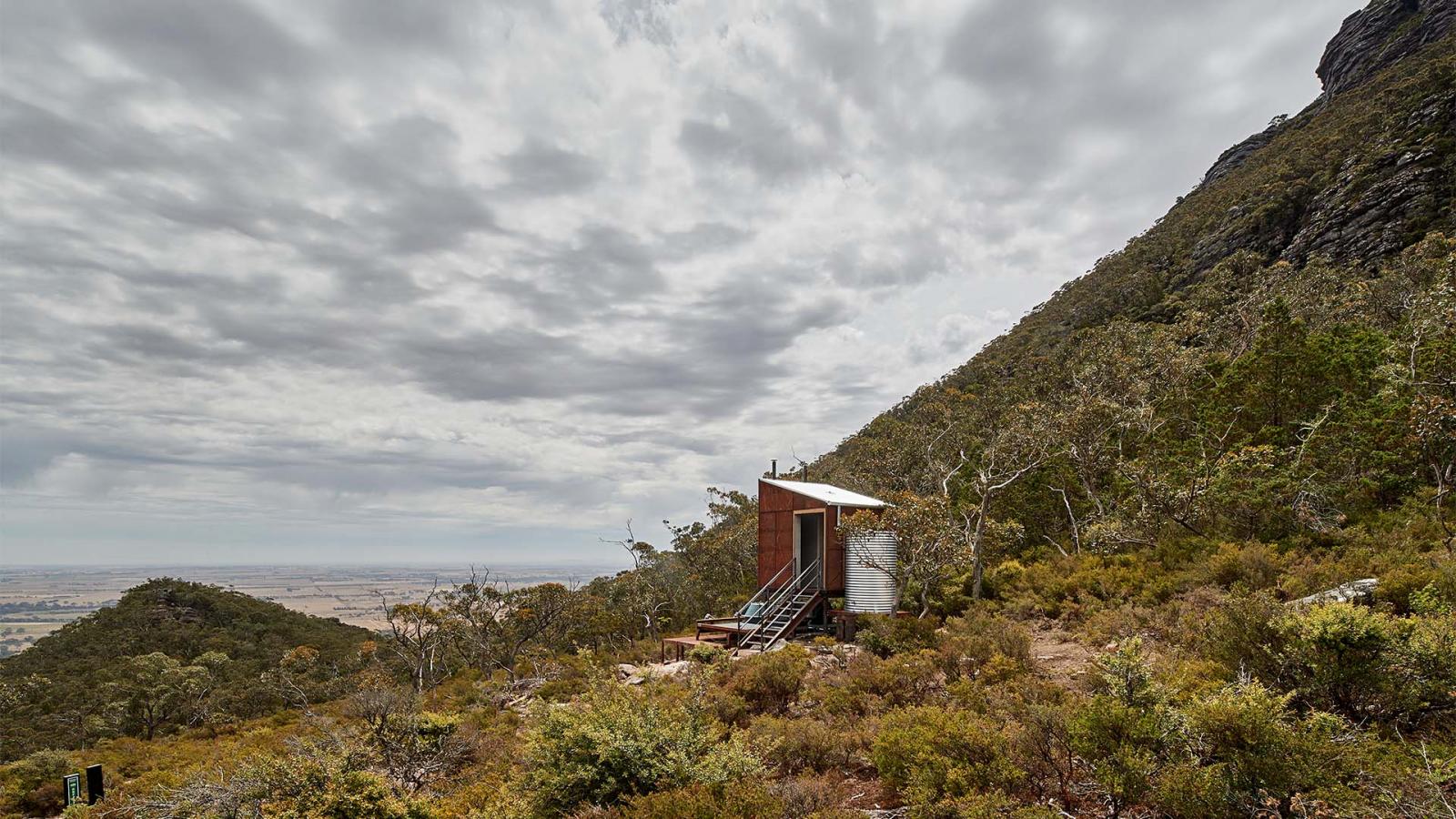 A small, rustic cabin with a water tank and solar panel sits on a hill amidst dense greenery along the Grampians Peaks Trail. The landscape slopes downward, leading to a distant flat expanse under a cloudy sky. A rocky mountain in Gariwerd rises to the right of the cabin.