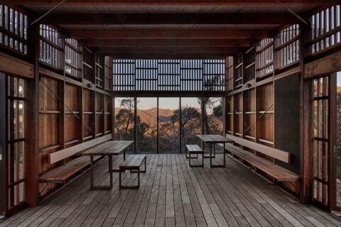 A wooden structure with open walls and a slatted roof, framed by vertical beams. Inside are two wooden picnic tables with attached benches. The background reveals a scenic view of the Grampians Peaks Trail in Gariwerd, showcasing a forested landscape and mountain range under a clear sky.