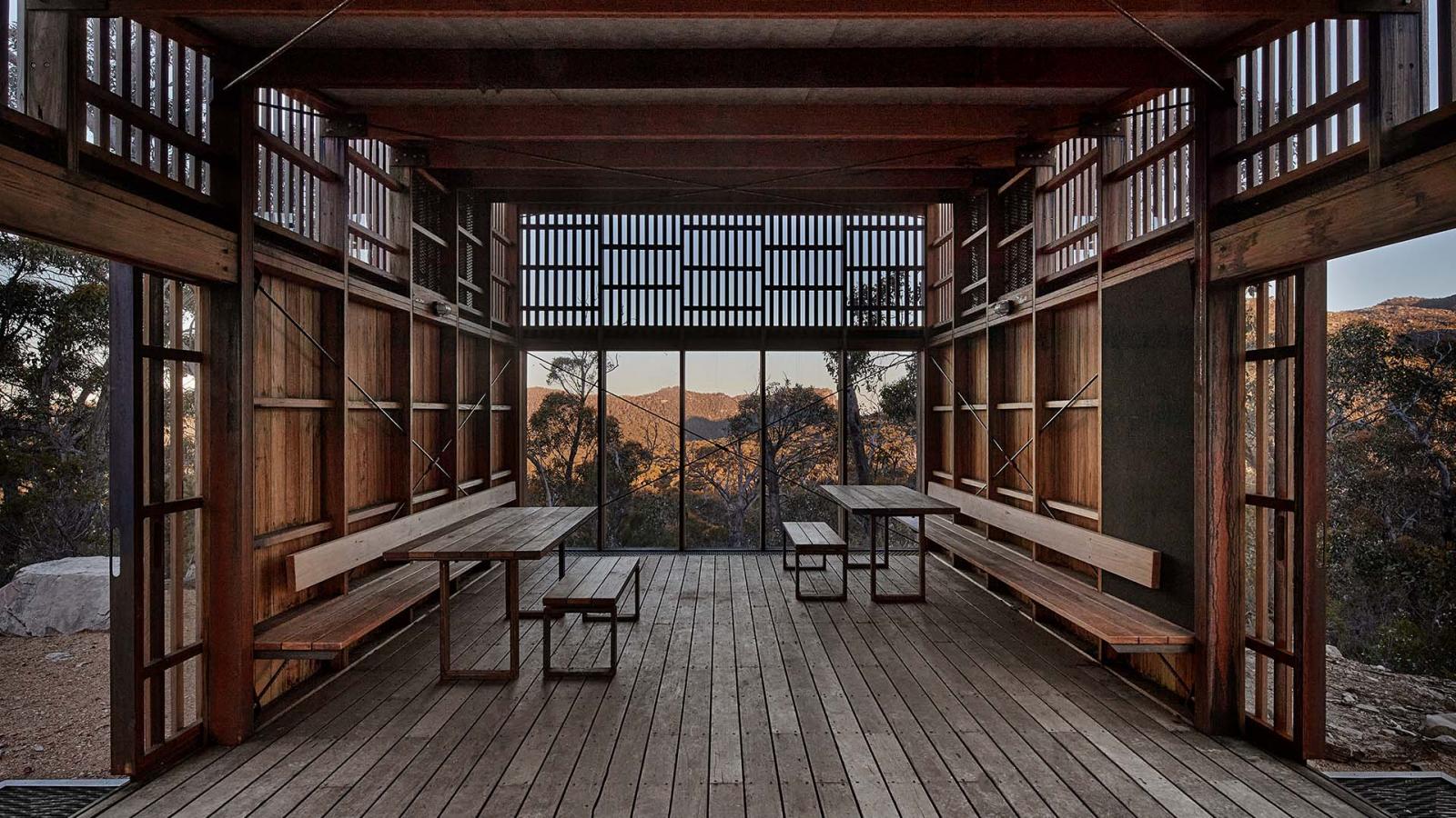 A wooden structure with open walls and a slatted roof, framed by vertical beams. Inside are two wooden picnic tables with attached benches. The background reveals a scenic view of the Grampians Peaks Trail in Gariwerd, showcasing a forested landscape and mountain range under a clear sky.