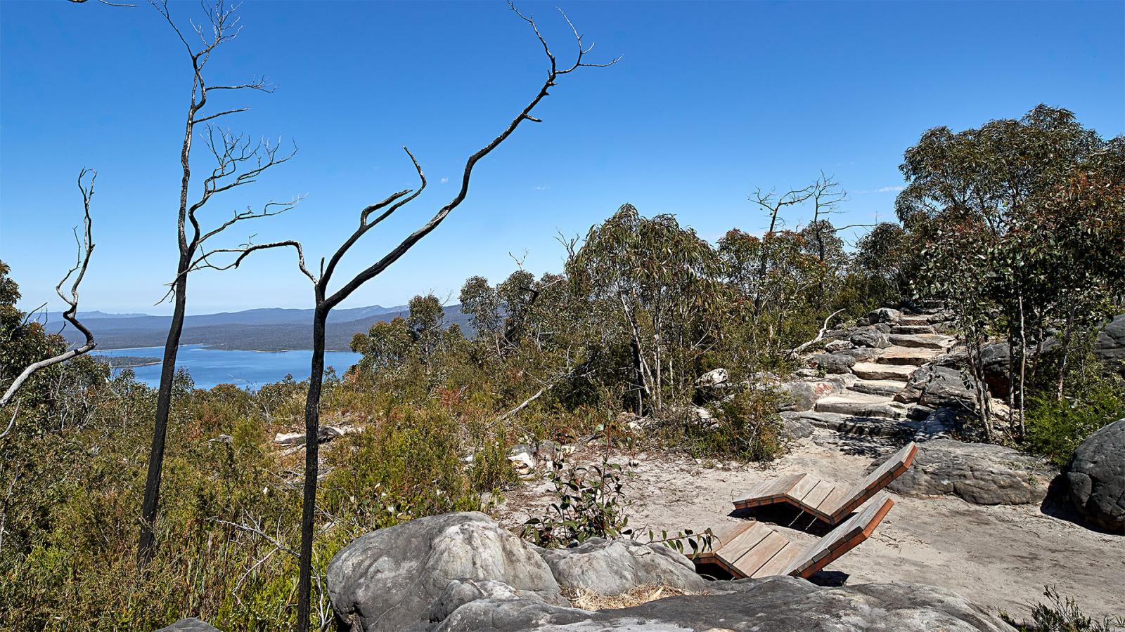 A scenic view of the Grampians Peaks Trail on a hilltop with a wooden bench and steps leading upwards. The area, part of Gariwerd, is surrounded by rocks and trees with sparse foliage, overlooking a distant body of water and mountain range under a clear blue sky.