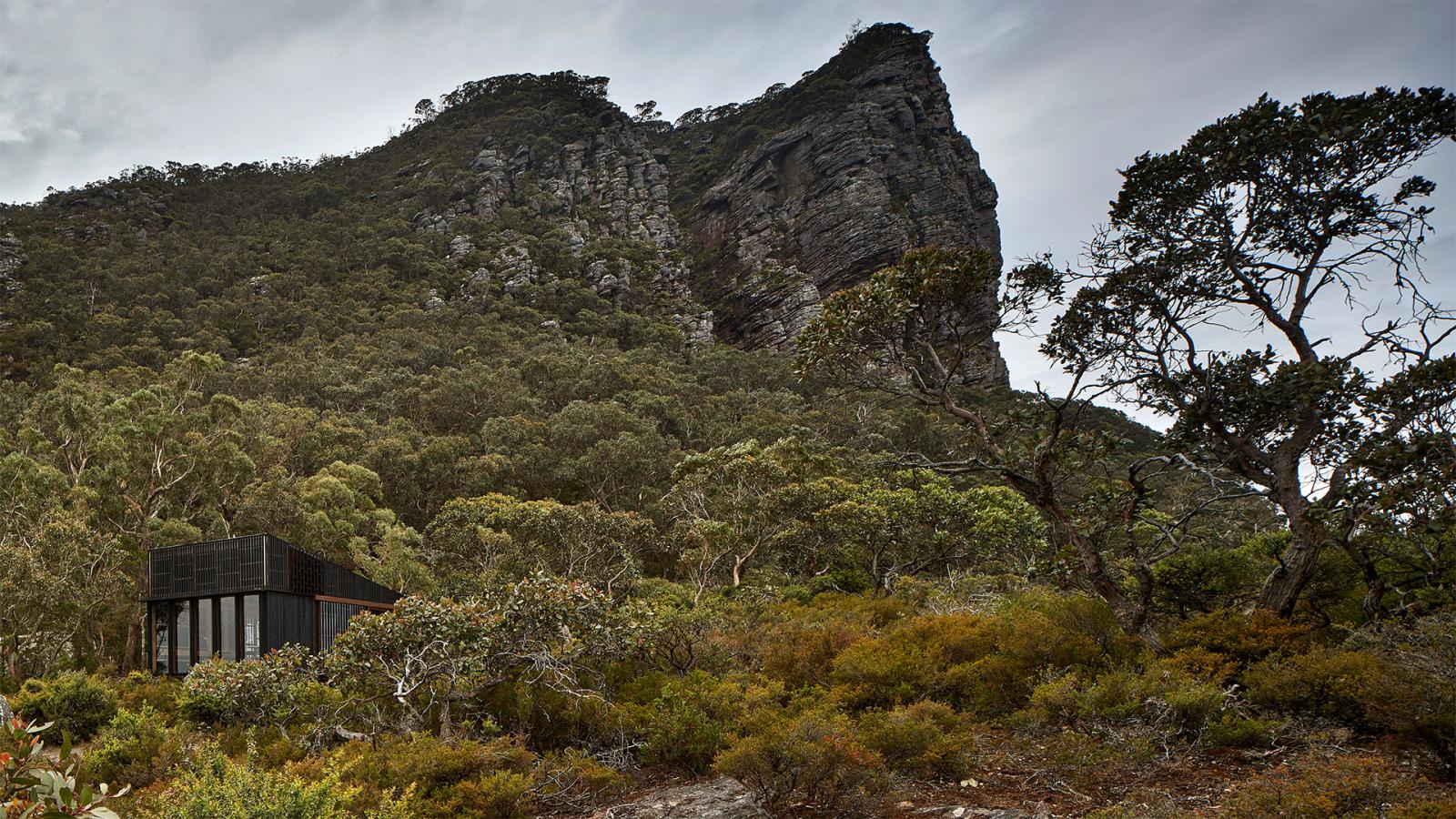 A small, modern black house sits at the base of Gariwerd, surrounded by dense, green forest. The rugged terrain consists of bushes, trees, and rocky ground under a cloudy sky. The mountain towers in the background, creating a dramatic natural backdrop along the Grampians Peaks Trail.