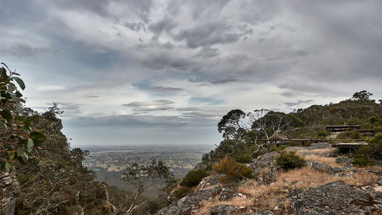 A scenic view from a mountaintop in Gariwerd, looking over a vast landscape with fields, trees, and distant horizon under a cloudy sky. To the right, along the Grampians Peaks Trail, there is a lookout structure built into the rocky terrain, surrounded by sparse vegetation.