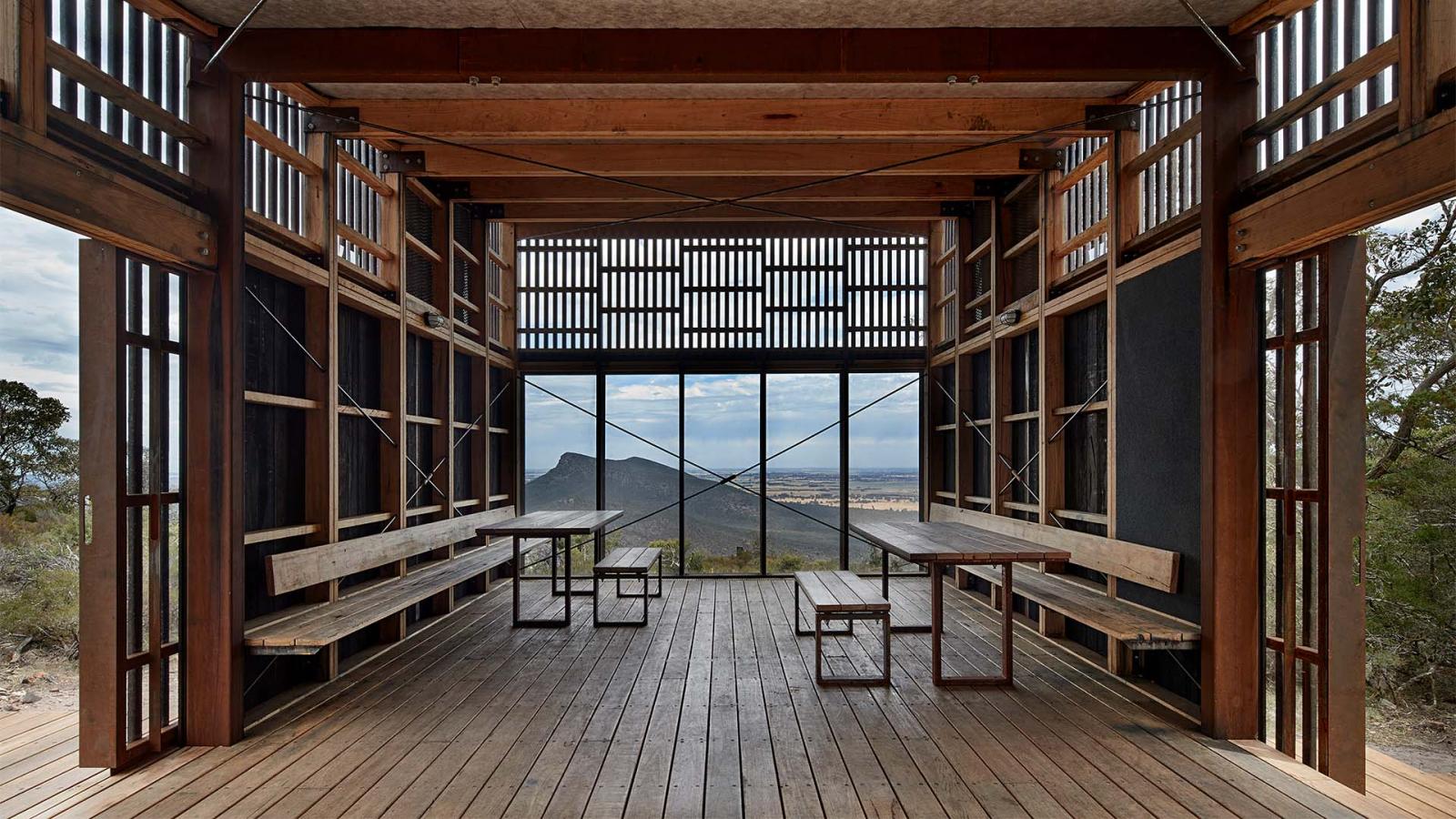 A rustic, open-air wooden pavilion with a panoramic view of the Gariwerd mountainous landscape in the distance. The interior features wooden benches and tables, and the walls have an open lattice design, allowing natural light to filter in. The setting is serene and inviting for those exploring the Grampians Peaks Trail.