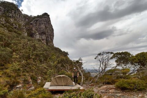In a rugged, mountainous landscape under a cloudy sky, a person stands next to a small pitched tent on a wooden platform. The scene is surrounded by dense vegetation and features a towering rock formation on the left, perfectly capturing the essence of the Grampians Peaks Trail in Gariwerd.