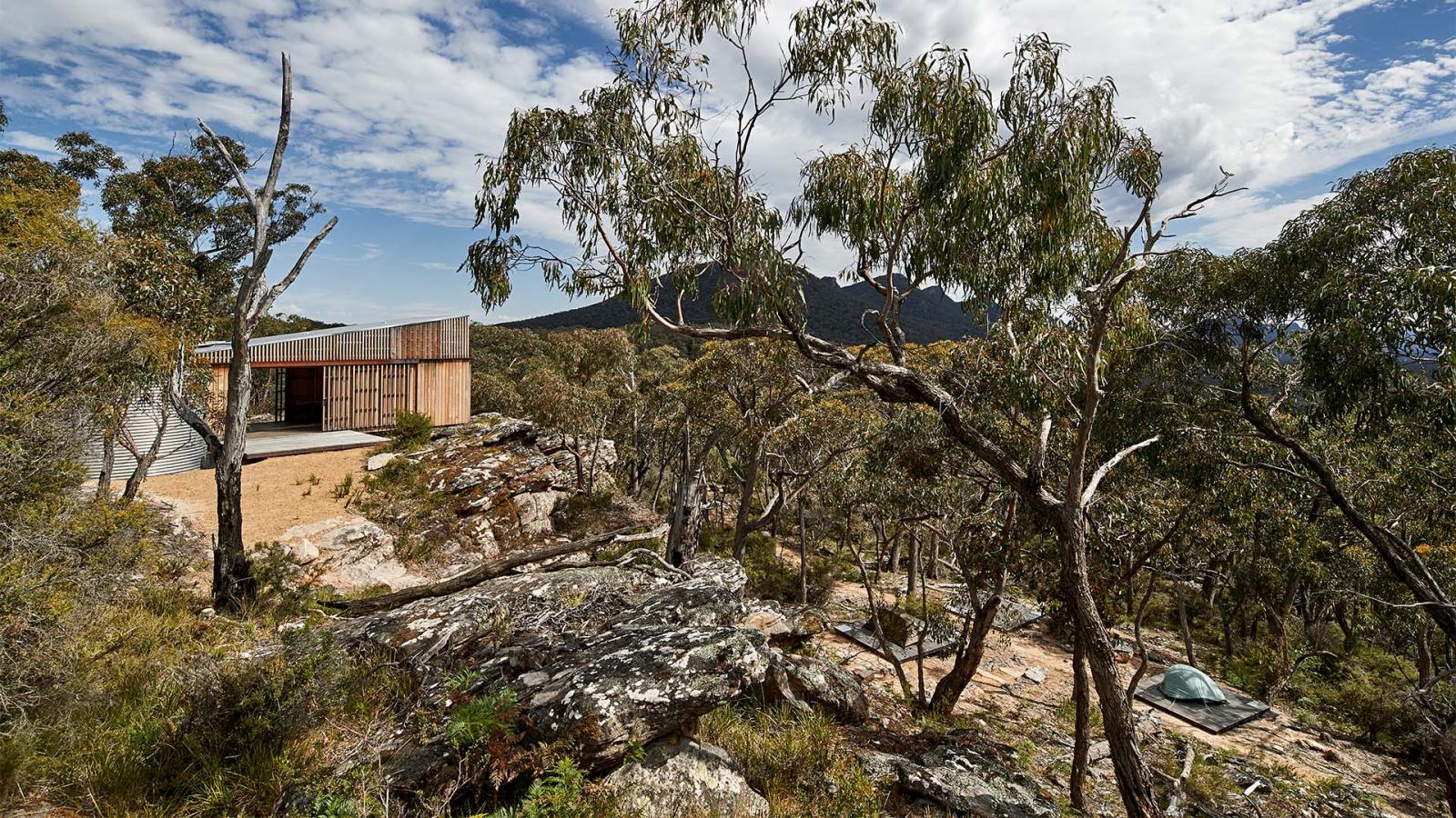 A modern house with a wooden exterior is perched on a rocky hill surrounded by dense trees. The sky is partly cloudy, with mountainous landscapes reminiscent of Gariwerd visible in the background. A small tent is set up on the ground near the house, offering adventurers easy access to the Grampians Peaks Trail.