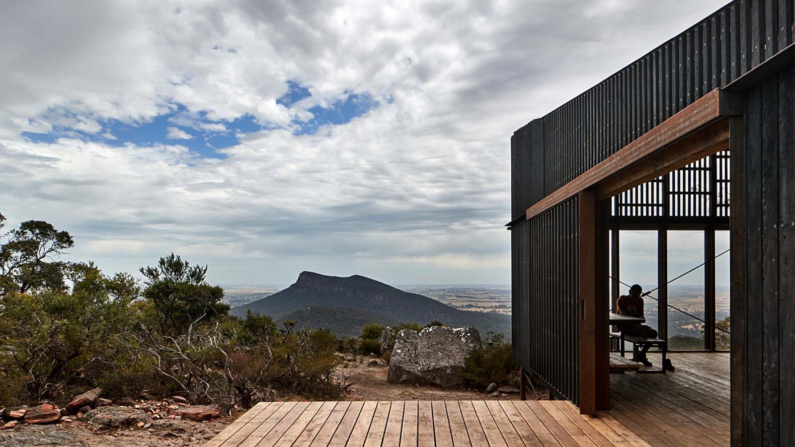 A modern, open-sided shelter with wooden flooring and a black structure overlooks a scenic landscape in Gariwerd. A person sits inside the shelter, while in the background, a distant mountain and expansive sky along the Grampians Peaks Trail add to the peaceful ambiance. Bushes and rocks surround the foreground.