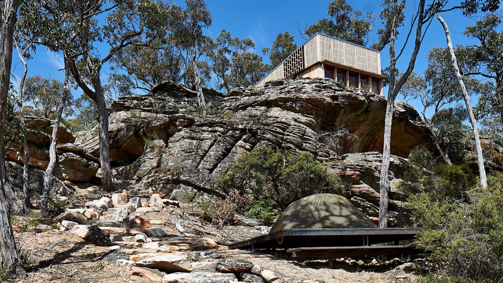 A modern house with large windows sits atop a rocky hillside surrounded by trees in the heart of Gariwerd. The rocky terrain features layered stone formations, and a wooden pathway, reminiscent of the Grampians Peaks Trail, leads up the hill. The structure beautifully integrates with the natural landscape.